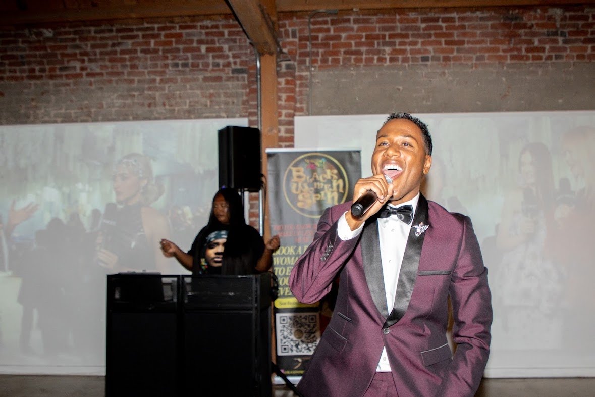 Aaron The Uplifter performing with a microphone in a burgundy tuxedo at the Black Beat Magazine relaunch event.