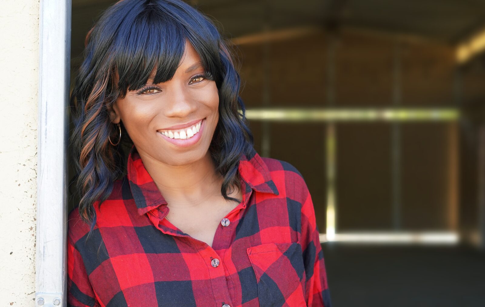 Angelique Bates smiling broadly in a red and black plaid shirt, leaning against a doorway.
