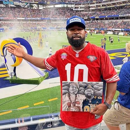Eric "E-Wash" Washington at SoFi Stadium, holding a framed piece of artwork at a football game.