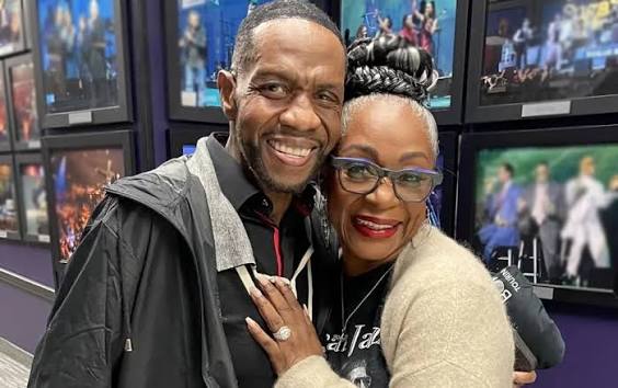 Freddie Jackson smiling and embracing Regina Belle in front of a wall of framed concert photographs.