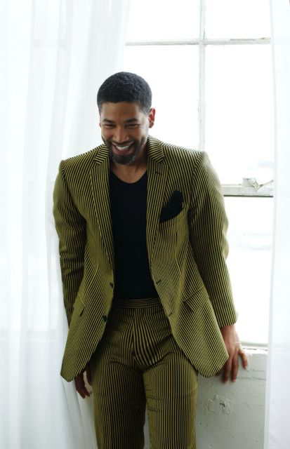 Jussie Smollett smiling, dressed in a green pinstripe suit, photographed against a bright window.