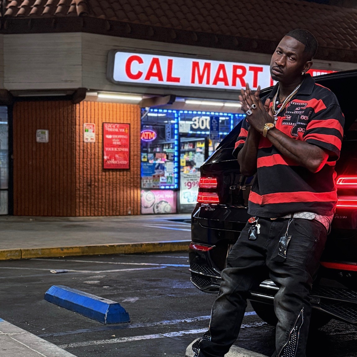 Krizko leaning against a black SUV at night outside the Cal Mart storefront in a red and black striped polo
