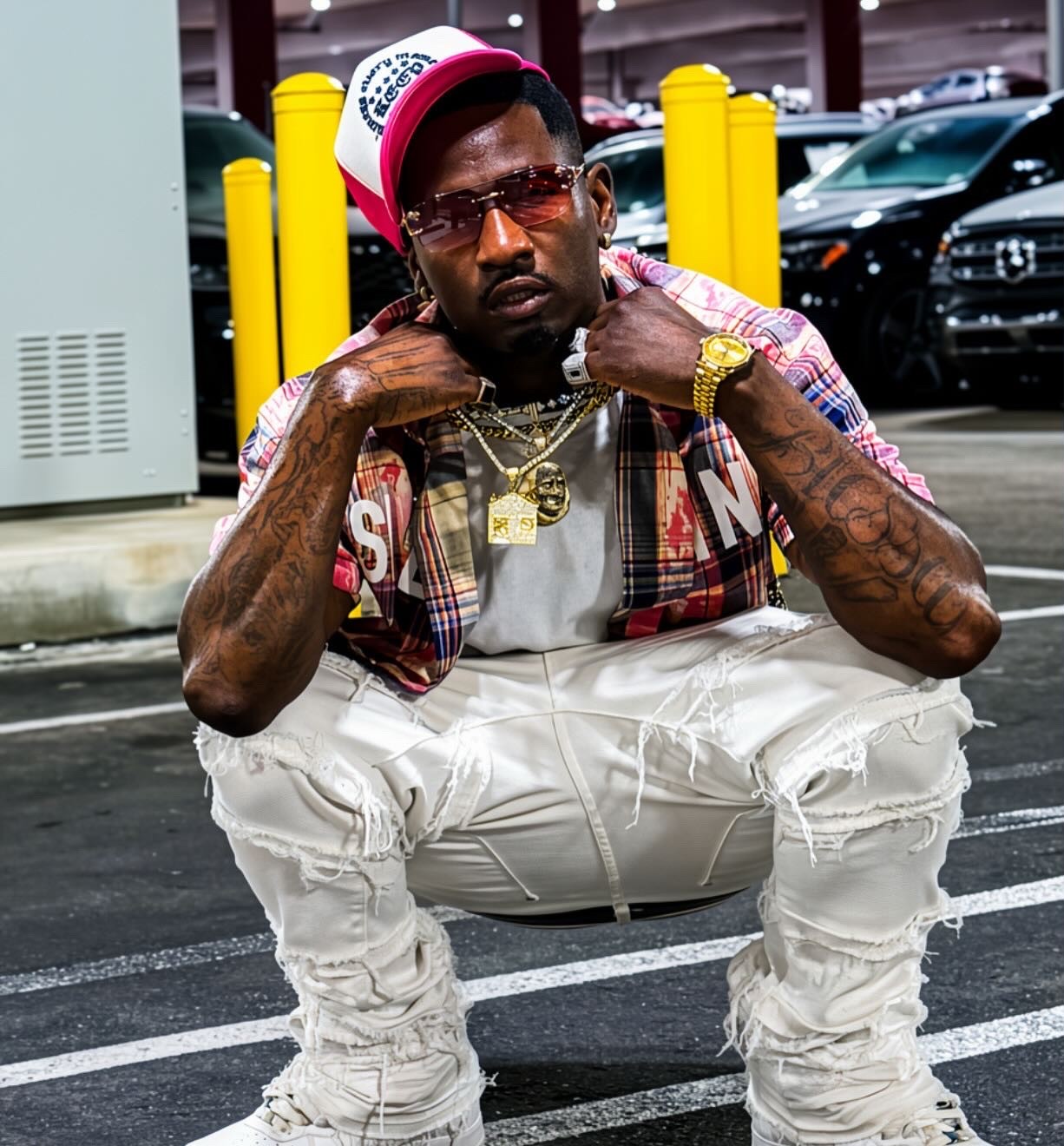 Krizko crouching in a parking structure wearing a pink and white trucker cap, tinted shades, plaid shirt and cream stacked jeans.
