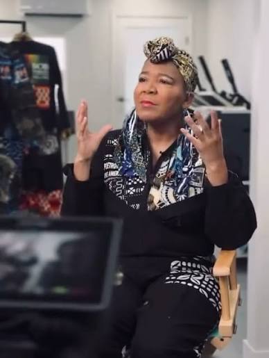 Roni Walter, PoetRoniGirl, seated in her studio in a black-and-white printed outfit and a patterned head wrap, gesturing with both hands mid-conversation.