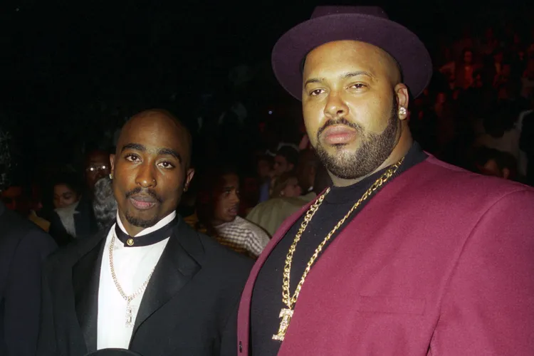 Tupac Shakur in a dark suit standing beside Death Row Records executive Suge Knight, who wears a maroon suit and hat, at an event in the mid-1990s.