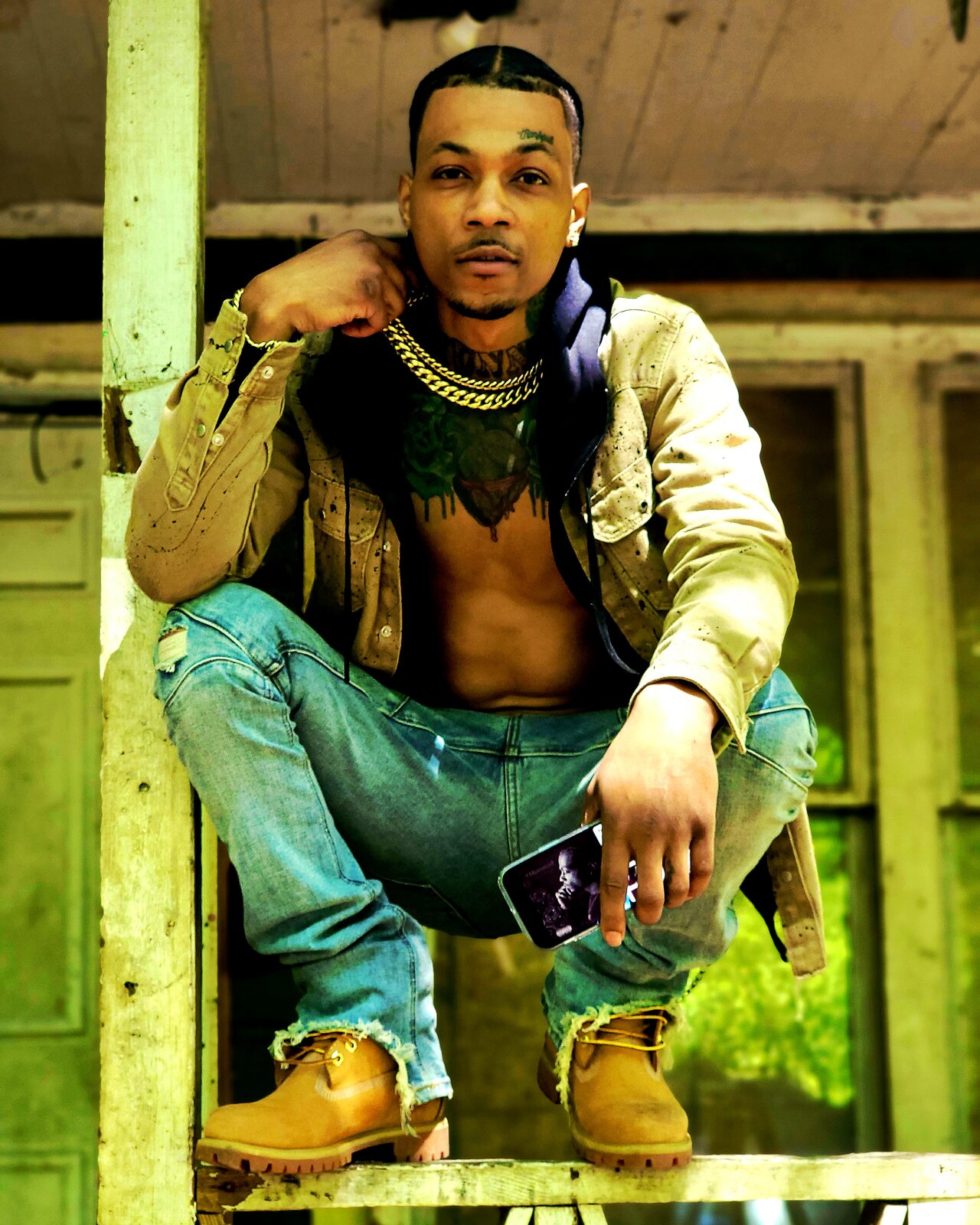 Wildlyfe 1Hunnud crouching on a weathered wooden porch railing in a tan jacket, gold chains and Timberland boots.