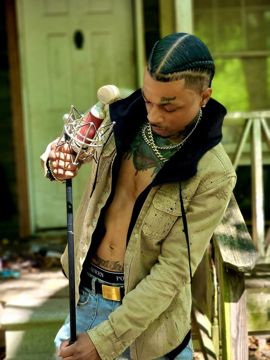 Wildlyfe 1Hunnud holding a studio microphone on its stand, wearing a tan splatter-print jacket and gold chains.