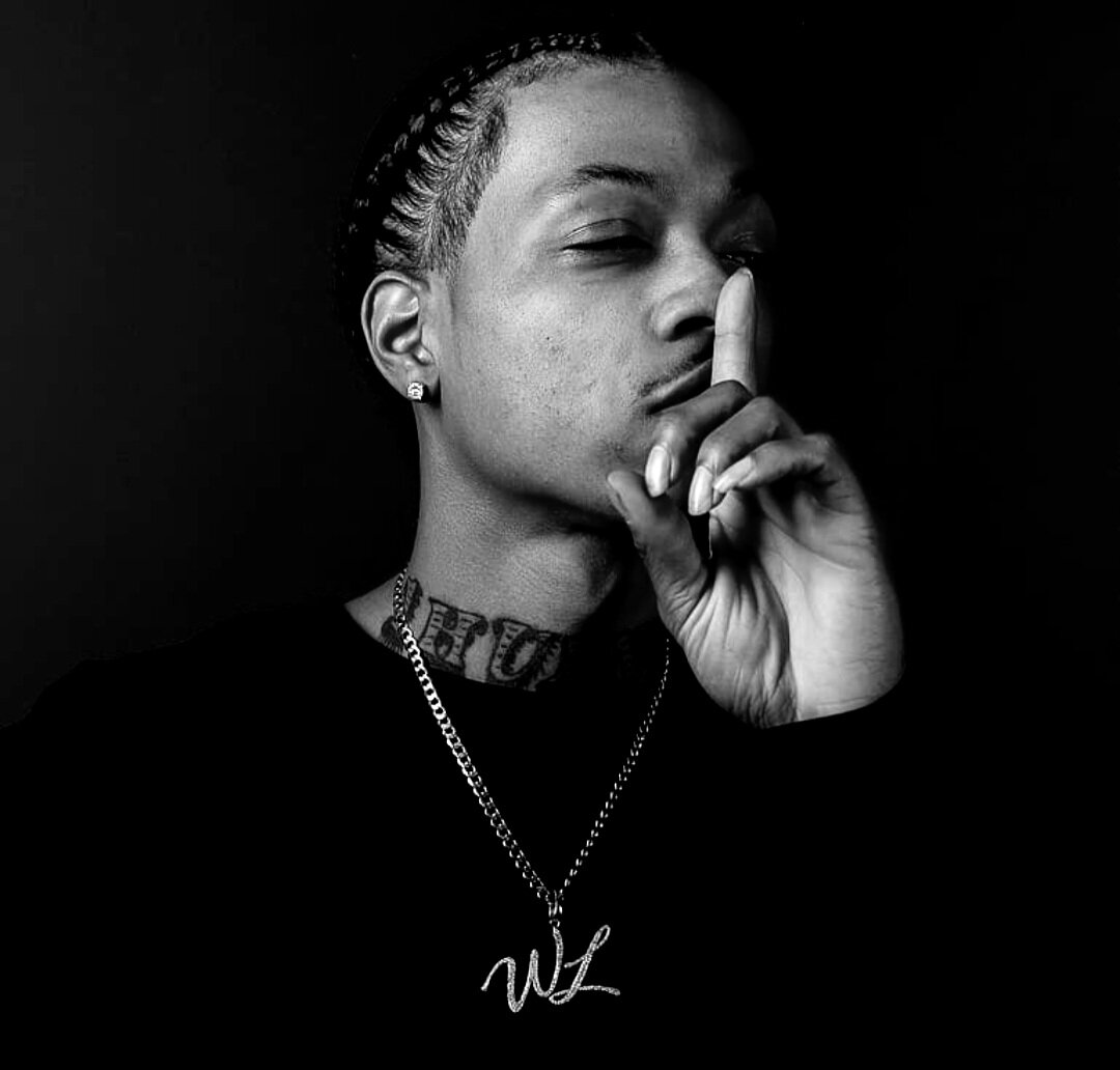 Black-and-white portrait of Wildlyfe 1Hunnud against a dark background, one finger raised to his lips, wearing a chain with a WL pendant.